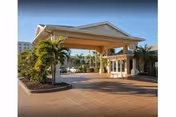 Entrance driveway with a large covered porte-cochère supported by columns at the Life Care Center of Melbourne, surrounded by palm trees and landscaped greenery under a clear sky.