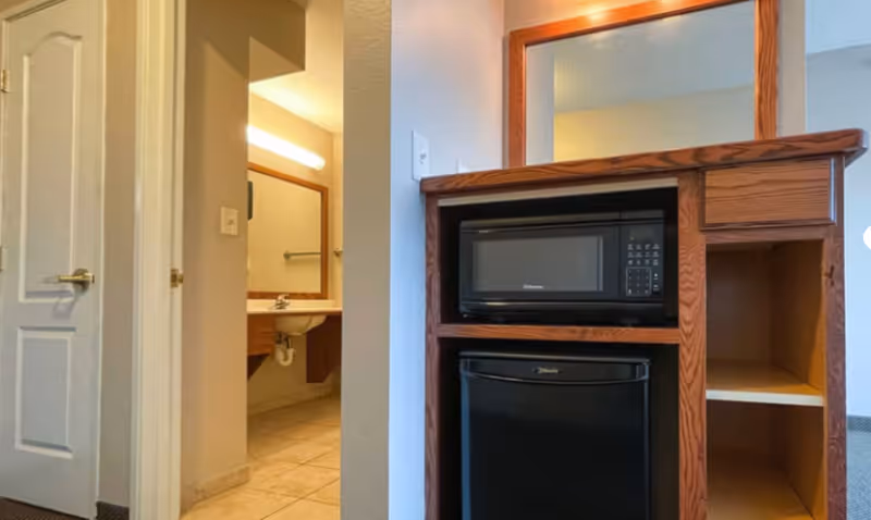 Interior view of a room in Lino Lakes Assisted Living showing a wooden cabinet with a microwave and mini refrigerator. To the left, there is a doorway leading to a bathroom with a sink, mirror, and tiled floor.