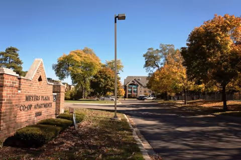 View of the entrance to Meyers Plaza Co-Op Apartments with a brick sign on the left and a paved driveway lined with trees showing autumn foliage. A multi-story building is visible in the background under a clear blue sky.