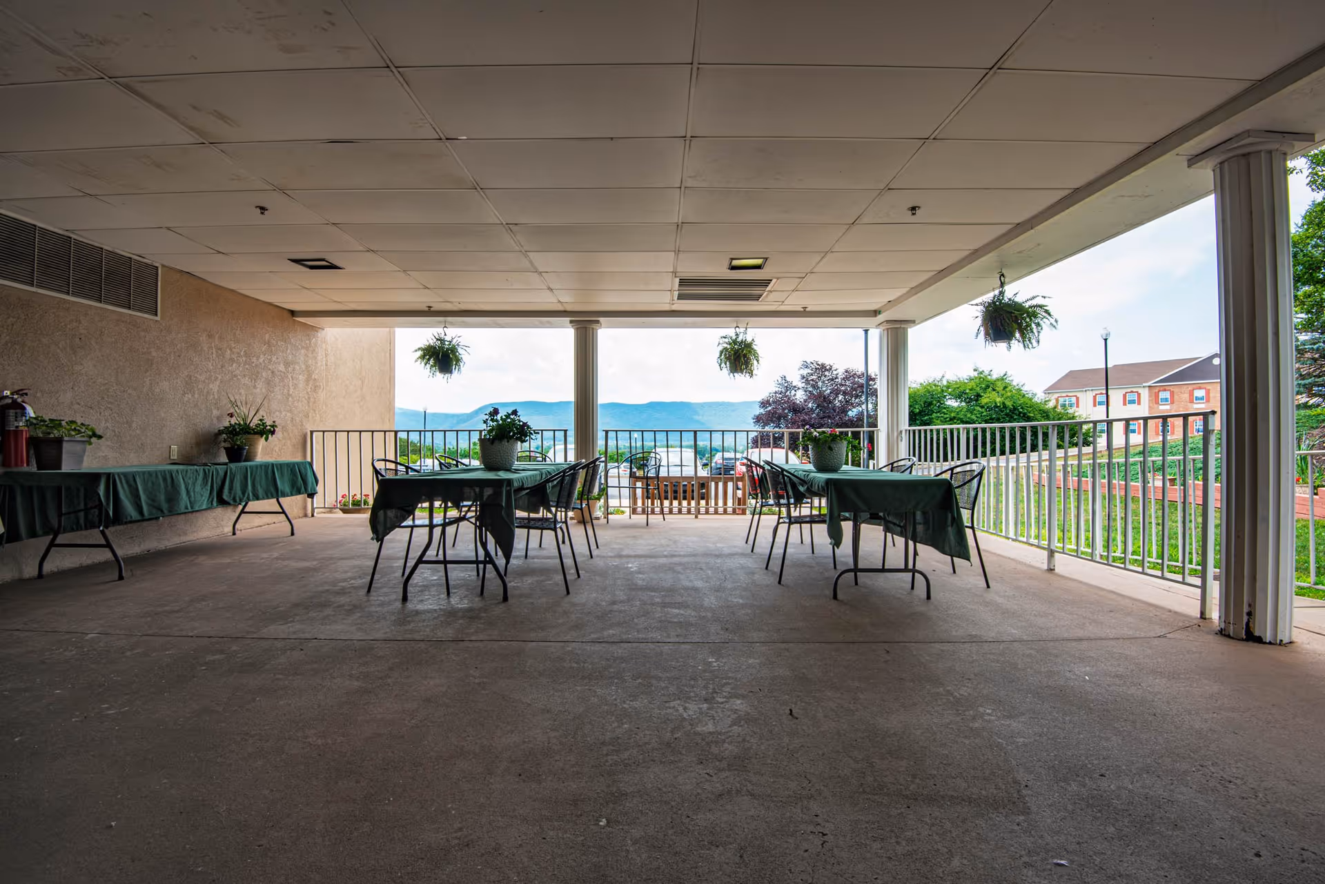 Covered outdoor patio area with tables and chairs arranged with green tablecloths and potted plants on top. Hanging plants are suspended from the ceiling. The patio overlooks a scenic view of distant hills and nearby buildings.