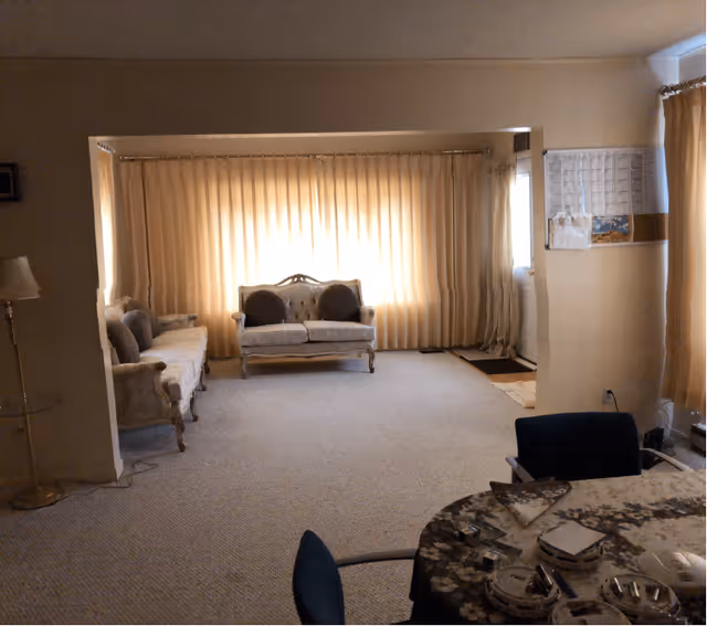 Living room with two vintage sofas facing a large window covered by beige curtains, carpeted floor, and a table in the foreground.