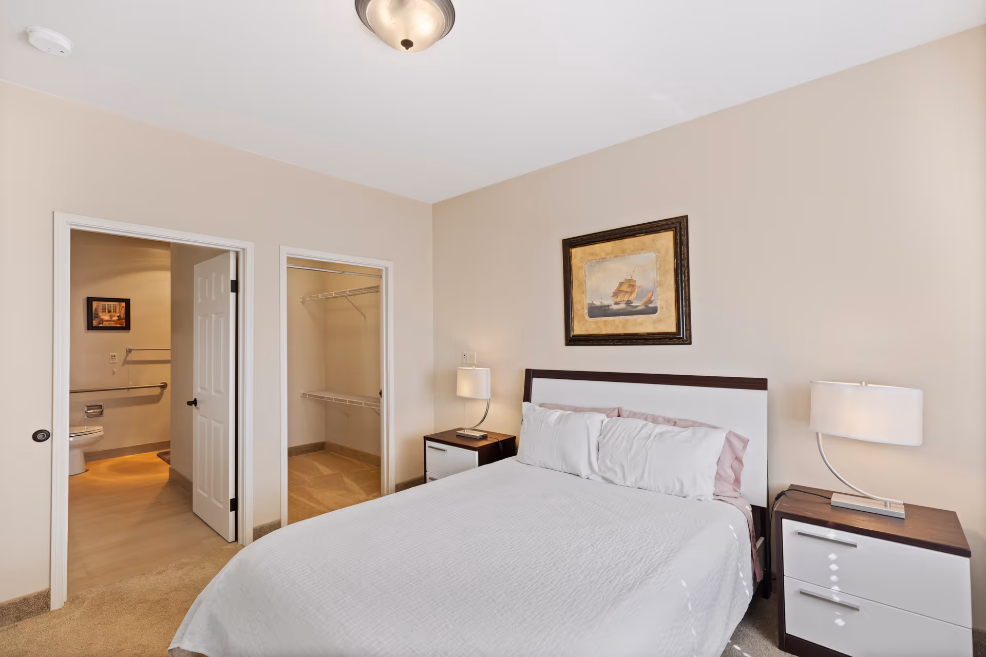 A tidy bedroom with a bed, two nightstands and lamps, a framed painting above the headboard, and open doors showing a bathroom and walk-in closet.