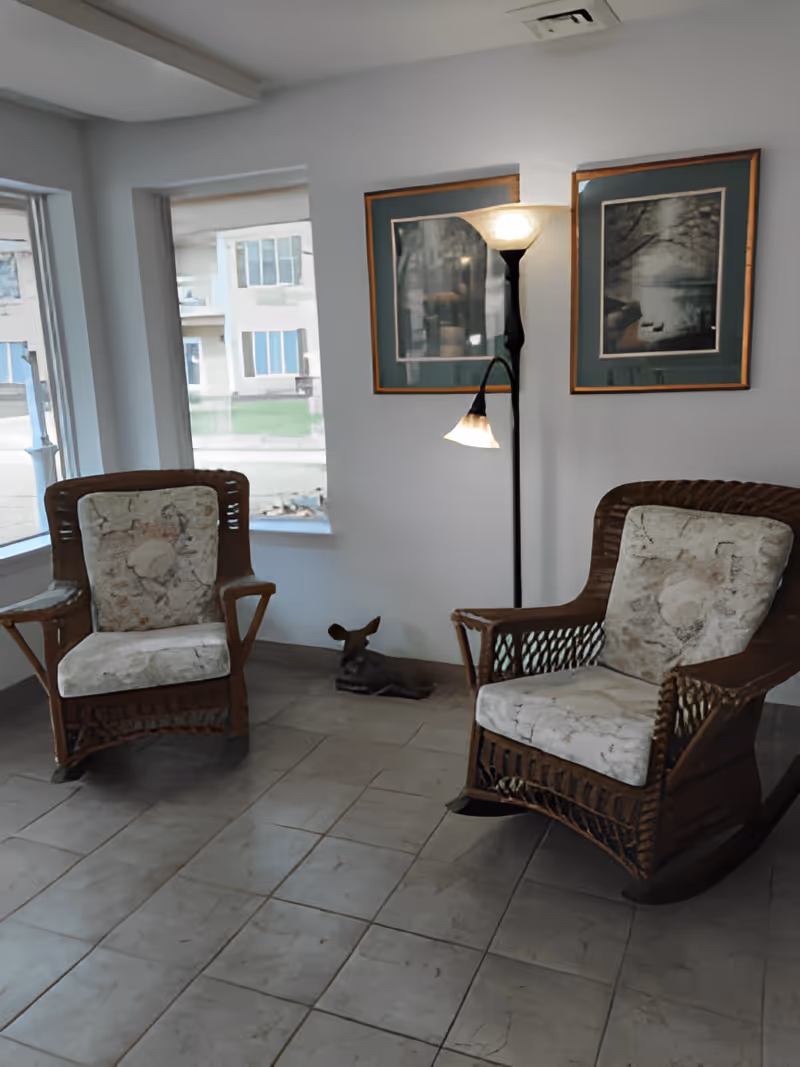 A cozy corner of a room with two cushioned wicker rocking chairs placed on a tiled floor. Between the chairs is a small decorative deer figurine on the floor. Behind the chairs, there is a tall floor lamp with two light fixtures, one at the top and one adjustable reading light. Two framed pictures with nature scenes hang on the wall above the lamp. Large windows let in natural light from the left side.