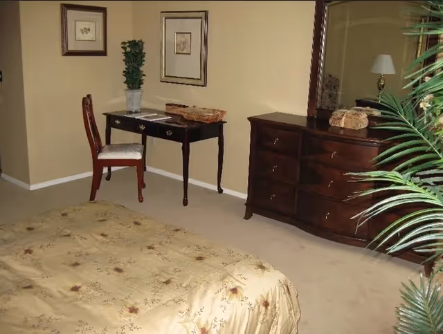 A bedroom with a beige floral bedspread, a dark wooden dresser with a large mirror, a small dark wooden desk with a chair, two framed pictures on the wall, a potted plant on the desk, and a table lamp partially visible behind a green plant in the foreground.
