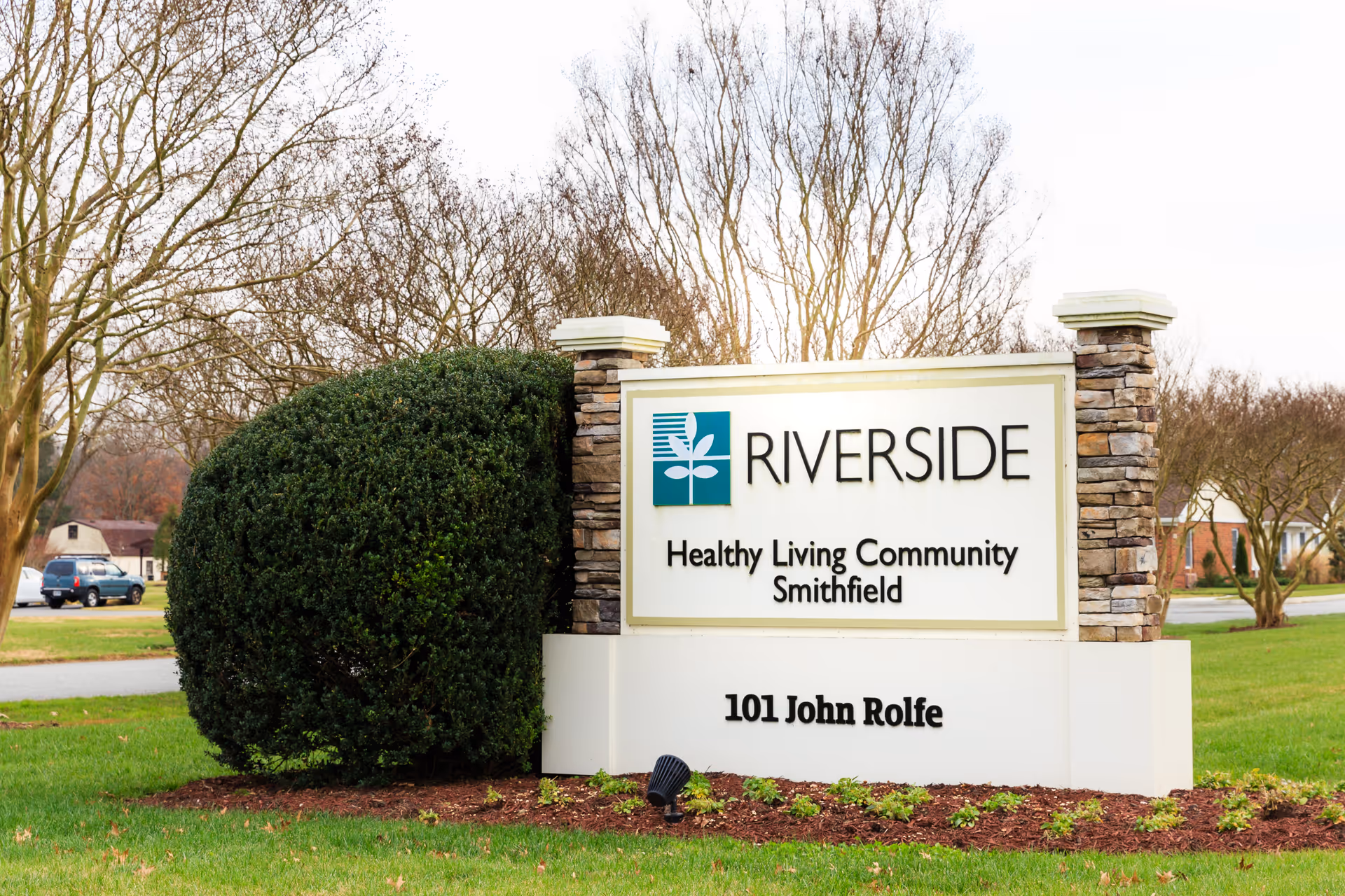 Outdoor view of a large stone and white sign for Riverside Healthy Living Community Smithfield, located at 101 John Rolfe, surrounded by grass, bushes, and leafless trees.