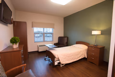 A tidy senior living bedroom with a single bed, bedside table and lamp, armchair, dresser with a potted plant, wall-mounted TV, and a window.