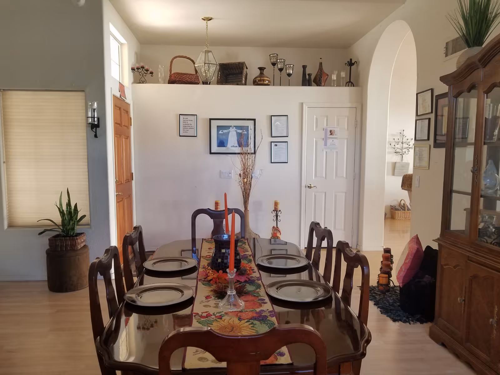 Sunlit dining room with a polished wooden table set for six, matching chairs, a china cabinet, and decorative wall accents.