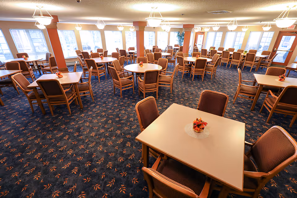 A spacious dining room with multiple square and round tables, each surrounded by four to six cushioned chairs. The tables have small floral centerpieces. The room has large windows along the walls letting in natural light, and the floor is covered with a patterned carpet. Ceiling lights provide additional illumination.