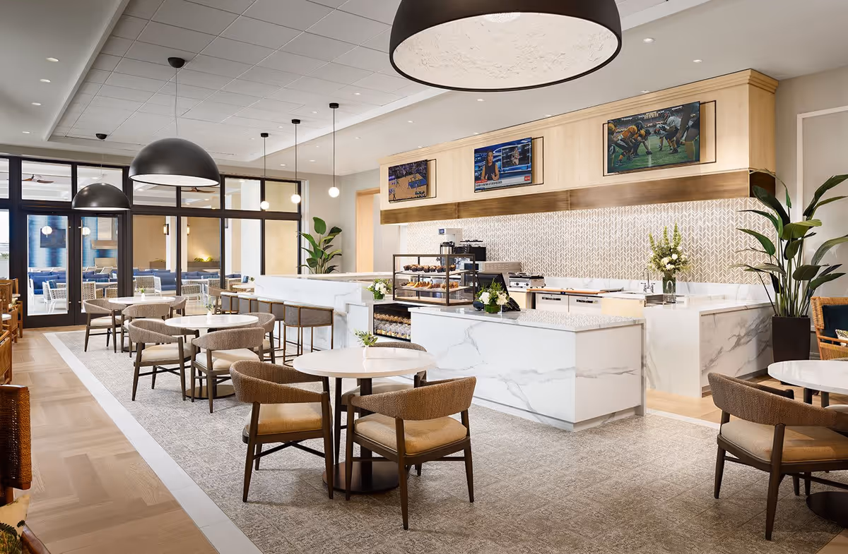 A modern dining area in a senior living facility featuring round tables with cushioned chairs, large black pendant lights, a marble counter with baked goods on display, and three wall-mounted televisions showing sports and news. The space has large windows and glass doors leading to another seating area.