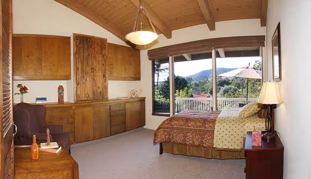 Spacious bedroom with a wooden vaulted ceiling, built-in cabinets, and a bed by large windows showing a patio and hillside view.