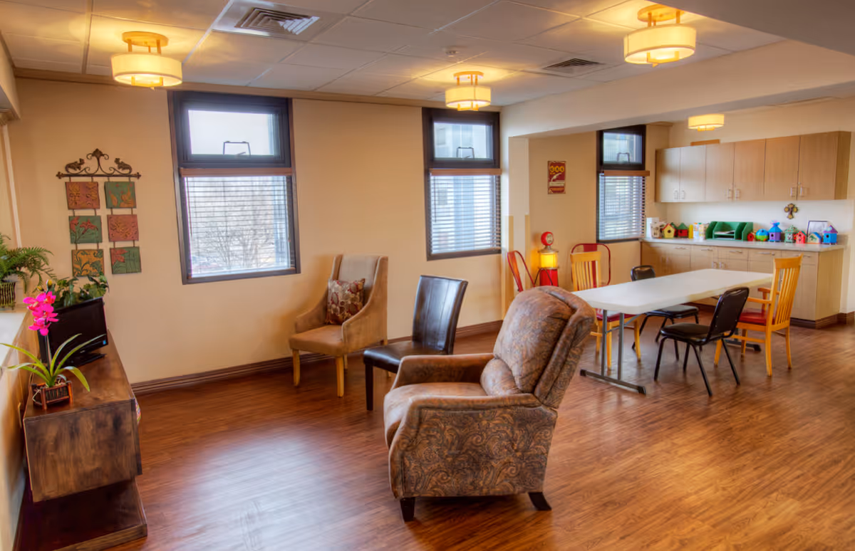 A well-lit common area with wooden flooring featuring a variety of chairs including an upholstered armchair, a cushioned chair, and wooden chairs around a white rectangular table. The room has three windows with blinds, ceiling lights, a wooden cabinet with plants and decorative items, and a countertop with colorful birdhouses and storage cabinets underneath.