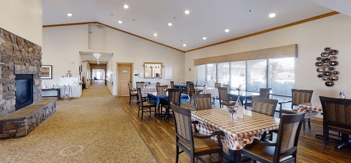 A dining room in an assisted living facility with several tables covered in checkered tablecloths, each decorated with small American flags and floral centerpieces. The room has large windows with blinds, wooden chairs, a stone fireplace on the left, and a hallway leading to other parts of the building.
