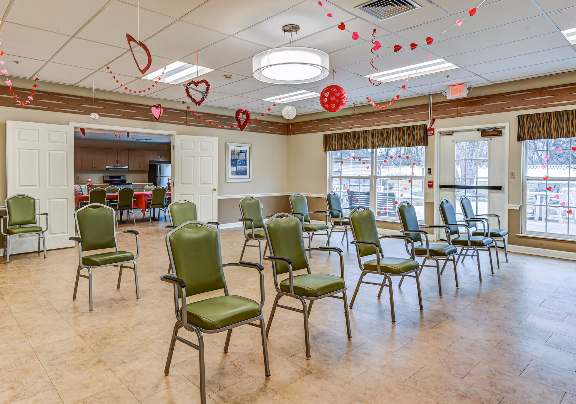 A spacious room with green cushioned chairs arranged in rows facing forward. The room has large windows with brown valances, letting in natural light. Red and pink heart-shaped decorations hang from the ceiling, suggesting a festive or Valentine's Day theme. Double doors open to an adjacent room with a kitchen and more chairs around a table. The floor is tiled, and the ceiling has recessed lighting and a circular ceiling light fixture.