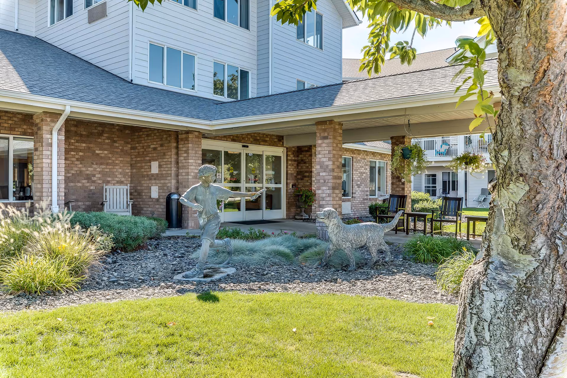 Outdoor view of Solstice Senior Living at Kennewick showing the entrance with brick pillars and sliding glass doors. In front of the entrance is a garden area with a statue of a boy playing with a dog. There are green plants, a tree on the right side, and outdoor seating with chairs and tables under a covered patio.
