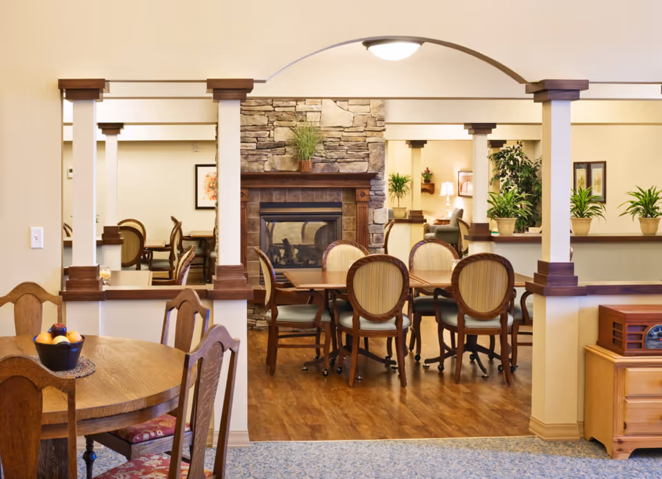 A cozy interior common area with wooden furniture including a round table with chairs and a rectangular table with six chairs on wheels. The room features a stone fireplace with a plant on the mantel, several potted plants, framed artwork on the walls, and warm lighting. The floor is a combination of wood and carpet, and the space is divided by white columns with wooden accents.