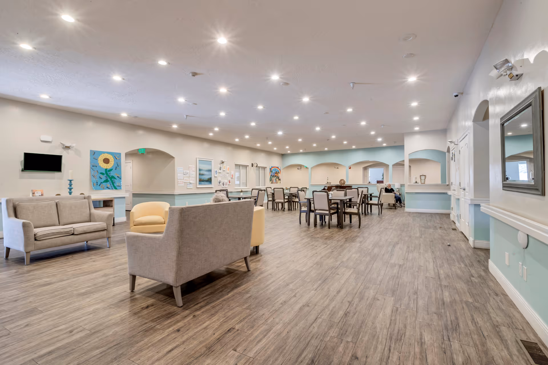 A spacious senior living facility common area with wooden flooring, multiple seating arrangements including sofas and chairs, several tables with chairs, soft lighting from ceiling fixtures, and pastel-colored walls adorned with artwork and a mirror.
