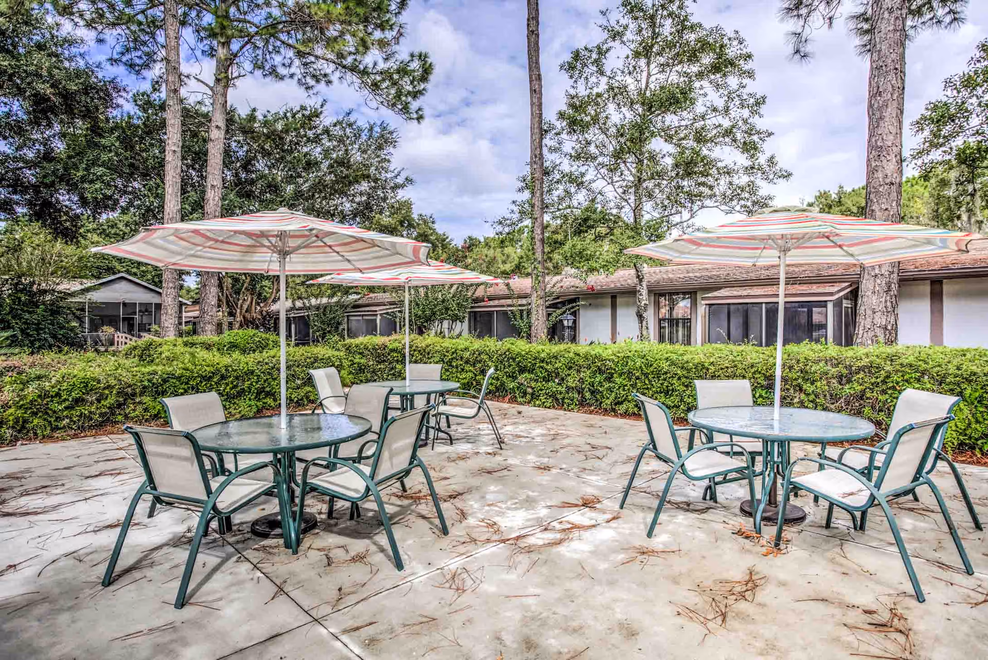 Outdoor patio area with two round glass tables, each surrounded by four chairs and shaded by large striped umbrellas. The patio is surrounded by green bushes and tall trees, with a building visible in the background under a partly cloudy sky.