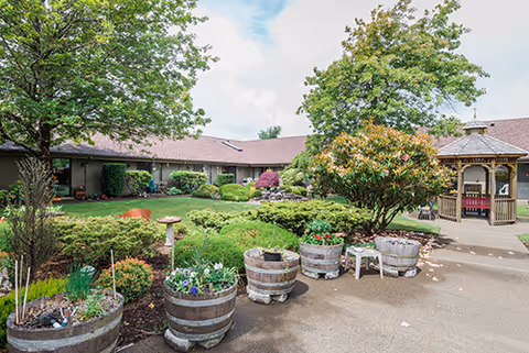 A landscaped outdoor courtyard at Channel Point Village featuring green shrubs, trees, and flower beds in wooden barrel planters. There is a paved walkway and a wooden gazebo on the right side. The building with a brown roof surrounds the courtyard under a partly cloudy sky.