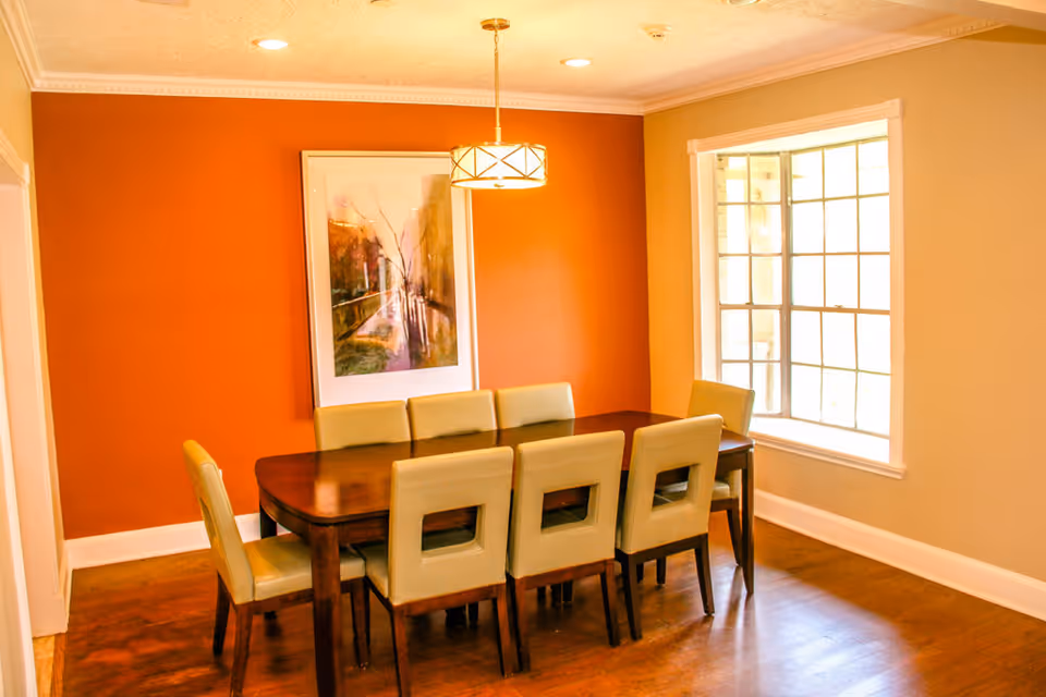 Warm dining room with orange accent wall, framed artwork, and beige upholstered chairs.