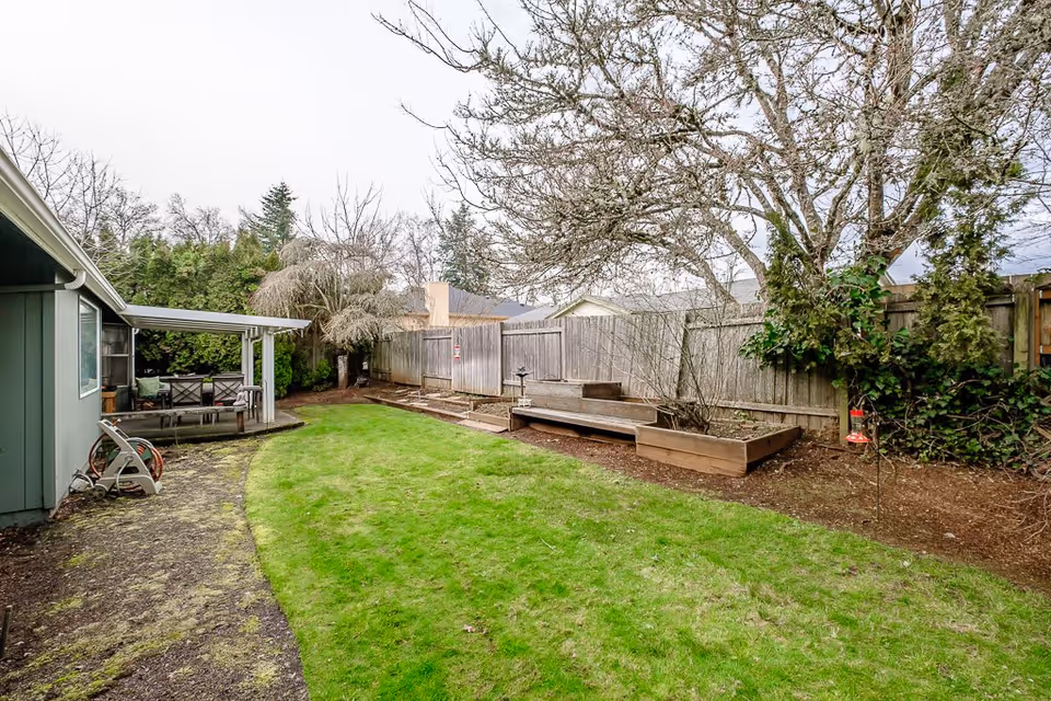 A backyard garden area with a green lawn, raised wooden garden beds along a wooden fence, leafless trees, and a covered patio with outdoor seating attached to a house.