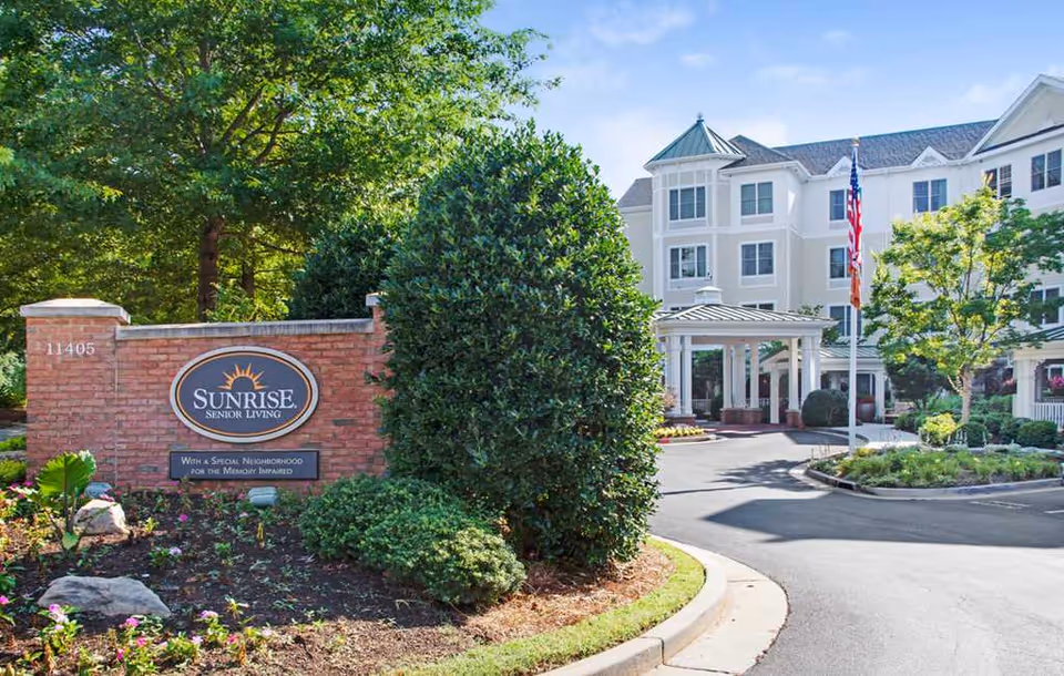 Entrance to Sunrise Senior Living facility with a brick sign displaying the Sunrise logo and text, surrounded by greenery and bushes. The multi-story building is visible in the background with a covered entrance, American flag, and landscaped driveway under a clear blue sky.