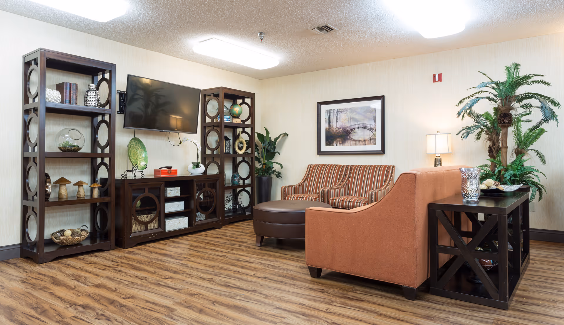 A furnished lounge with a sofa and loveseat, round ottoman, wall-mounted TV, decorative shelving, plants, and framed artwork on wood flooring.