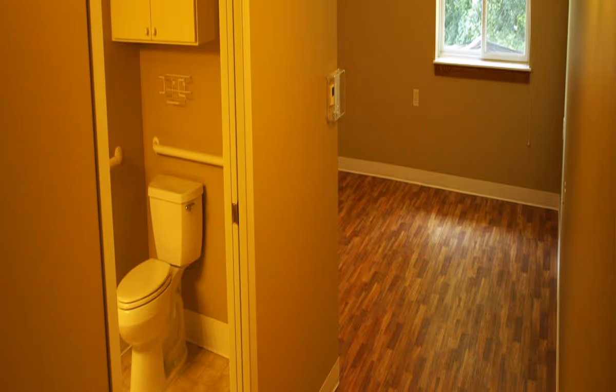Small interior showing a toilet in an open bathroom doorway leading into an empty room with wood-look flooring and a window.