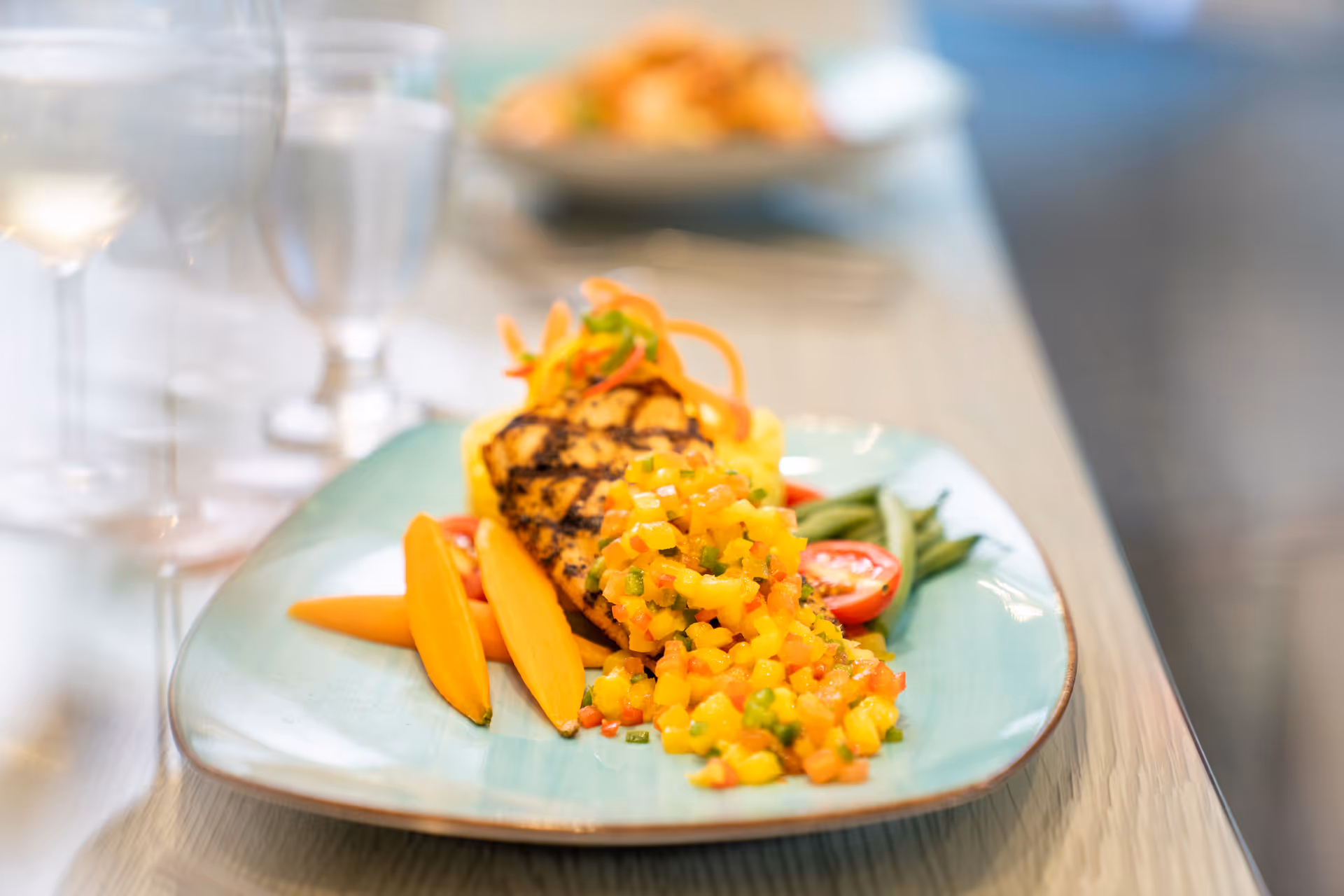 A plated grilled chicken breast topped with a colorful salsa and served with vegetables on a table with glassware.