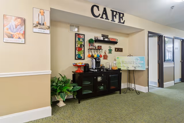 Interior hallway area of a senior living facility with a small cafe setup. The word 'CAFE' is displayed above a recessed area containing a black cabinet with a coffee maker, cups, and a juice dispenser. There are decorative wall hangings, a potted plant on the floor, and a sign on a stand next to the cabinet. The hallway has beige walls, green carpet, and doors leading to other rooms.