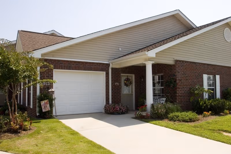 Exterior view of a single-story brick and siding residential building with a white garage door, a front porch with a column, a door decorated with a wreath, and landscaping including bushes and flowers.
