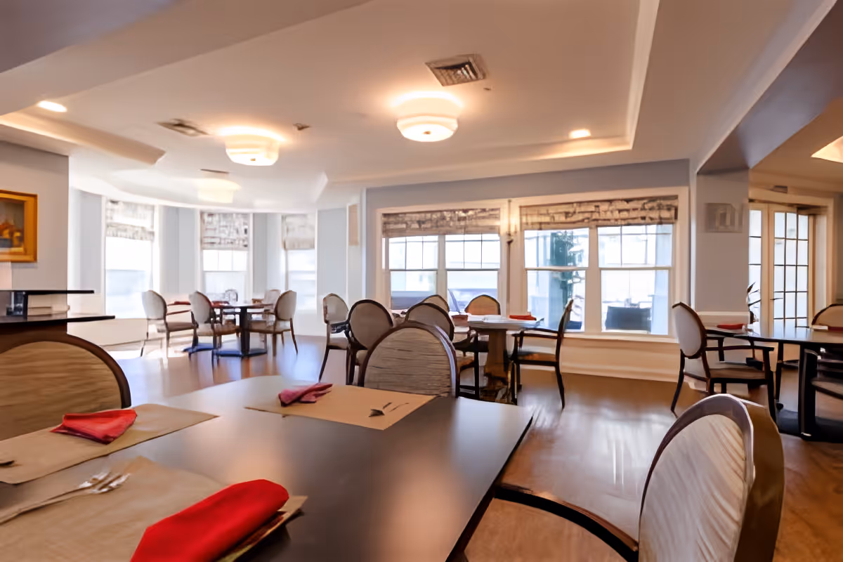 Dining room with multiple tables and chairs set with placemats and red napkins, large windows and ceiling lights.