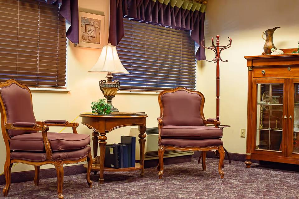 A cozy sitting area with two upholstered armchairs flanking a wooden side table topped with a lamp, beside a coat rack and glass-front cabinet.
