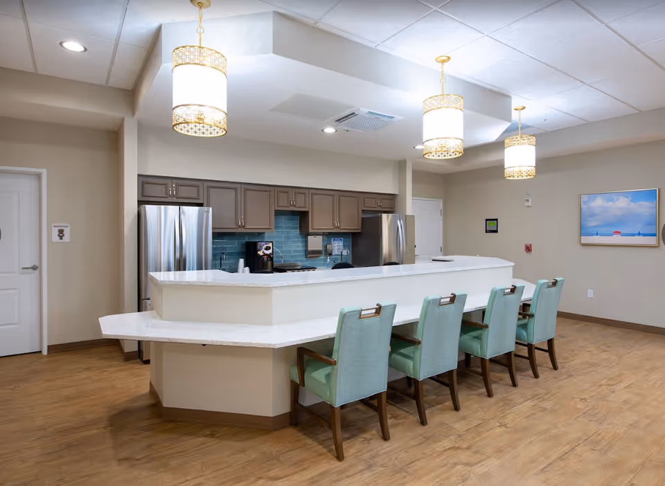 Bright communal kitchen and dining bar with five mint-green chairs at a white countertop, stainless steel appliances, and pendant lights.