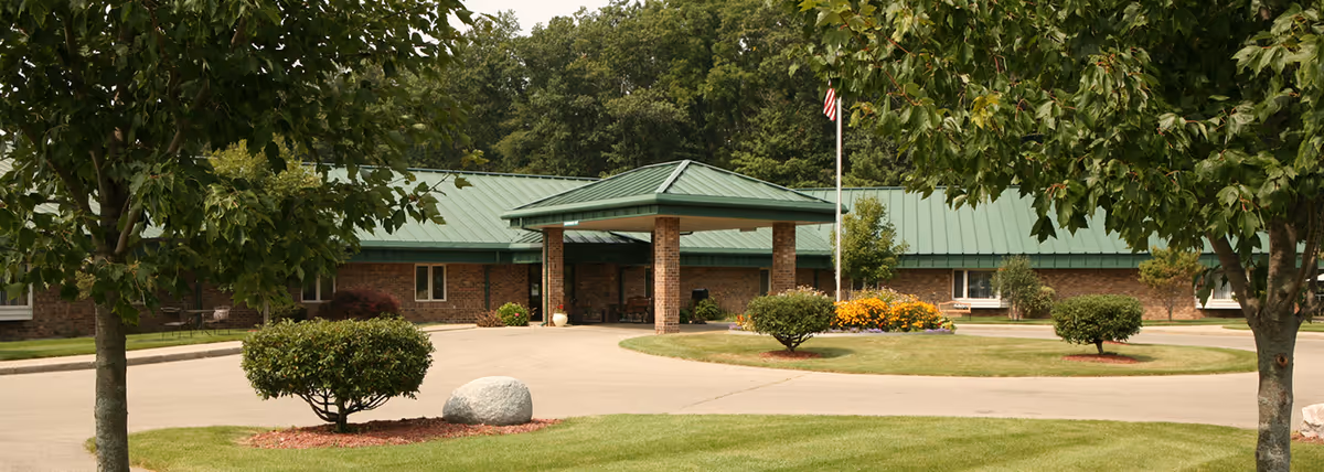 Front entrance of a single-story brick senior living building with a green metal roof, circular driveway, flagpole, and landscaped trees and shrubs.