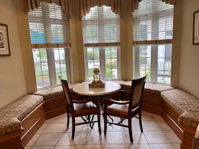 A cozy seating area with a round table and two wooden chairs with patterned cushions, surrounded by built-in cushioned benches along three large windows with white blinds and valances, overlooking a garden outside.