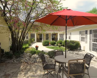 Outdoor courtyard area with a round table and four chairs under a large red umbrella. The courtyard is surrounded by a single-story building with multiple windows. There are bushes, a tree with pink blossoms, and a stone-covered garden bed along the walkway.