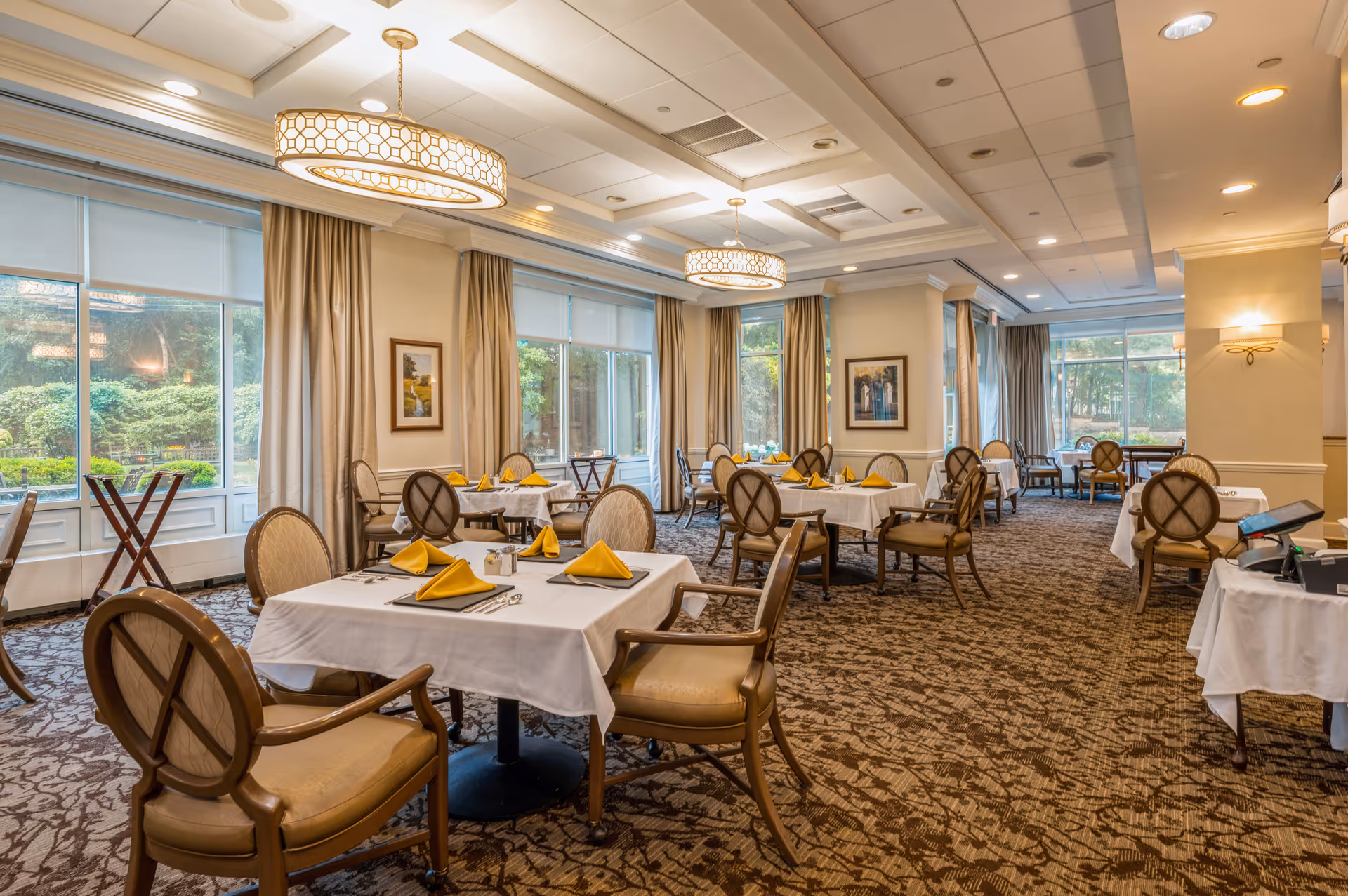 A spacious dining room with multiple tables covered in white tablecloths and set with yellow folded napkins, silverware, and small condiment containers. The room features large windows with beige curtains, patterned carpet, elegant ceiling lights, and framed artwork on the walls. The atmosphere is bright and inviting with natural light coming through the windows.