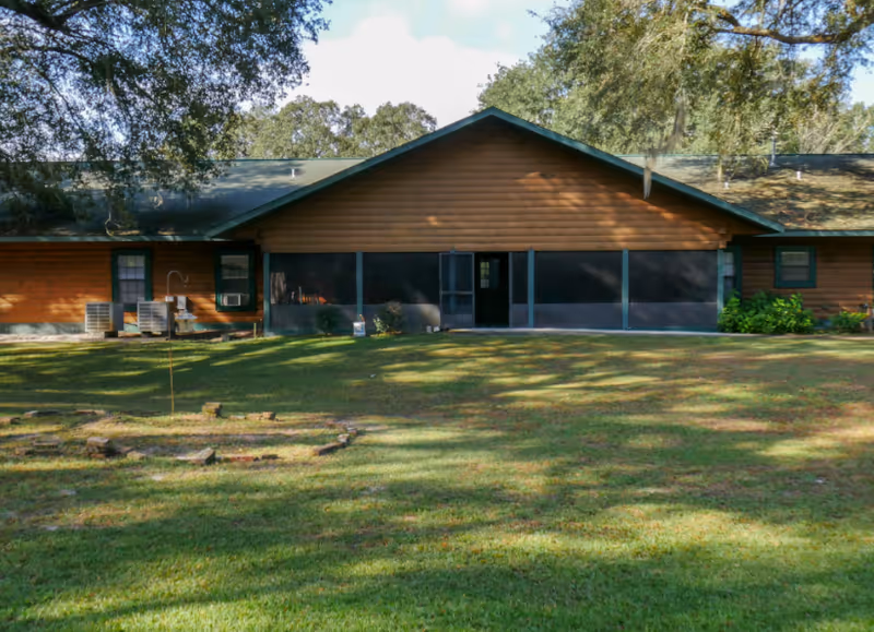 Single-story wood building with large dark screens, green lawn, and surrounding trees.