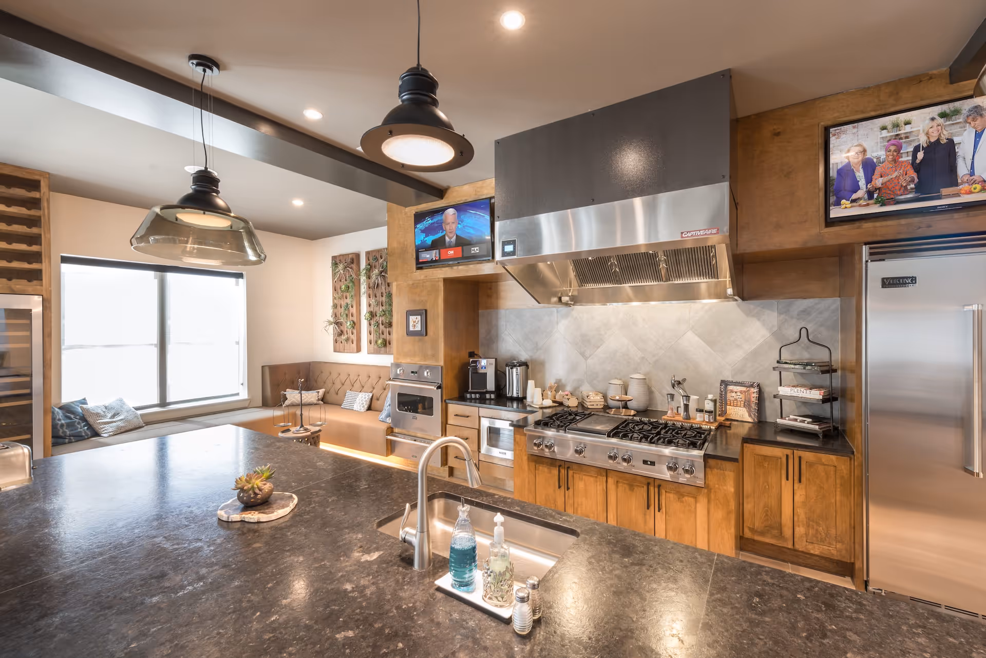 Modern kitchen area with a large dark countertop island featuring a sink with soap dispensers. The kitchen has wooden cabinets, a stainless steel stove with a large vent hood, a built-in oven, and a stainless steel refrigerator. There is a cozy seating nook with cushions by a large window, two wall-mounted TVs displaying news and a cooking show, and decorative wall art with plants.