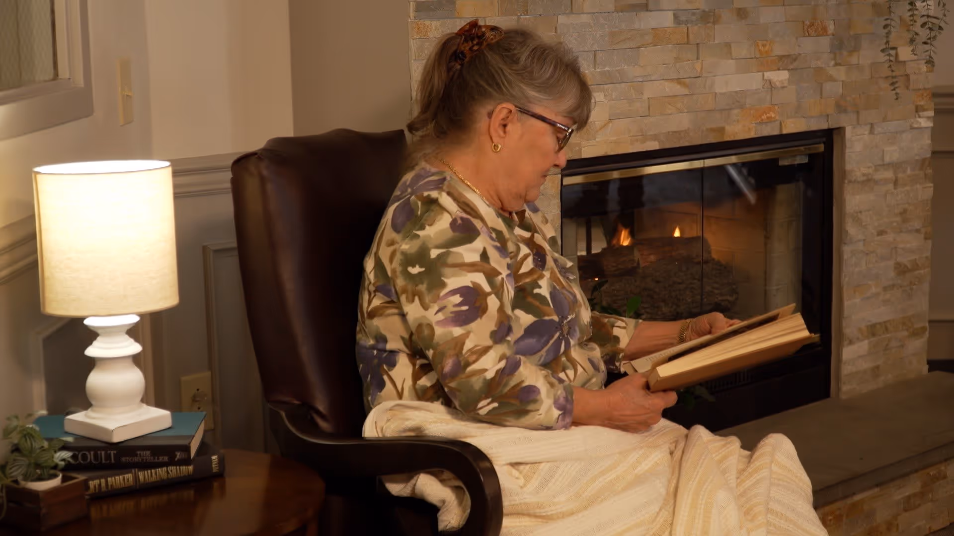 An elderly woman with glasses and a floral patterned shirt sits in a brown leather armchair, reading a book. She is covered with a light-colored blanket. Behind her is a stone fireplace with a fire burning. To her left, there is a wooden side table with a white lamp, some books, and a small potted plant.
