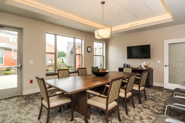 A modern meeting or dining room with a rectangular wooden table surrounded by eight cushioned chairs. The room has large windows letting in natural light, a wall-mounted flat-screen TV, a sideboard with decorative items, and a ceiling light fixture. The floor is carpeted with a patterned design, and there is a glass door leading outside.