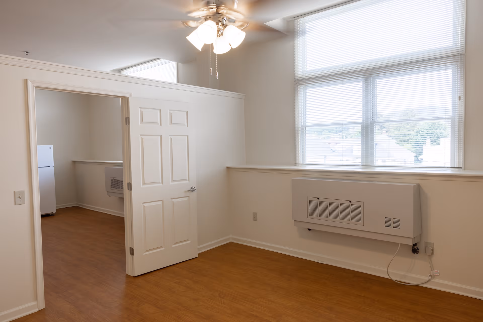 Empty room with wooden floor, white walls, a ceiling fan with lights, a large window with blinds, and a wall-mounted heater. An open door leads to another room with a white refrigerator visible.