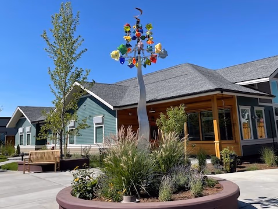 Outdoor courtyard area of a senior living facility with a modern sculpture featuring colorful glass pieces on a tall metal pole, surrounded by plants and flowers in a circular planter. There are benches, trees, and a building with green siding and large windows under a clear blue sky.