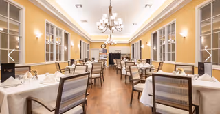 A spacious dining room with multiple tables covered in white tablecloths, each set with napkins, glasses, and small floral centerpieces. The room features large windows on both sides, warm yellow walls, elegant chandeliers, and wooden flooring.