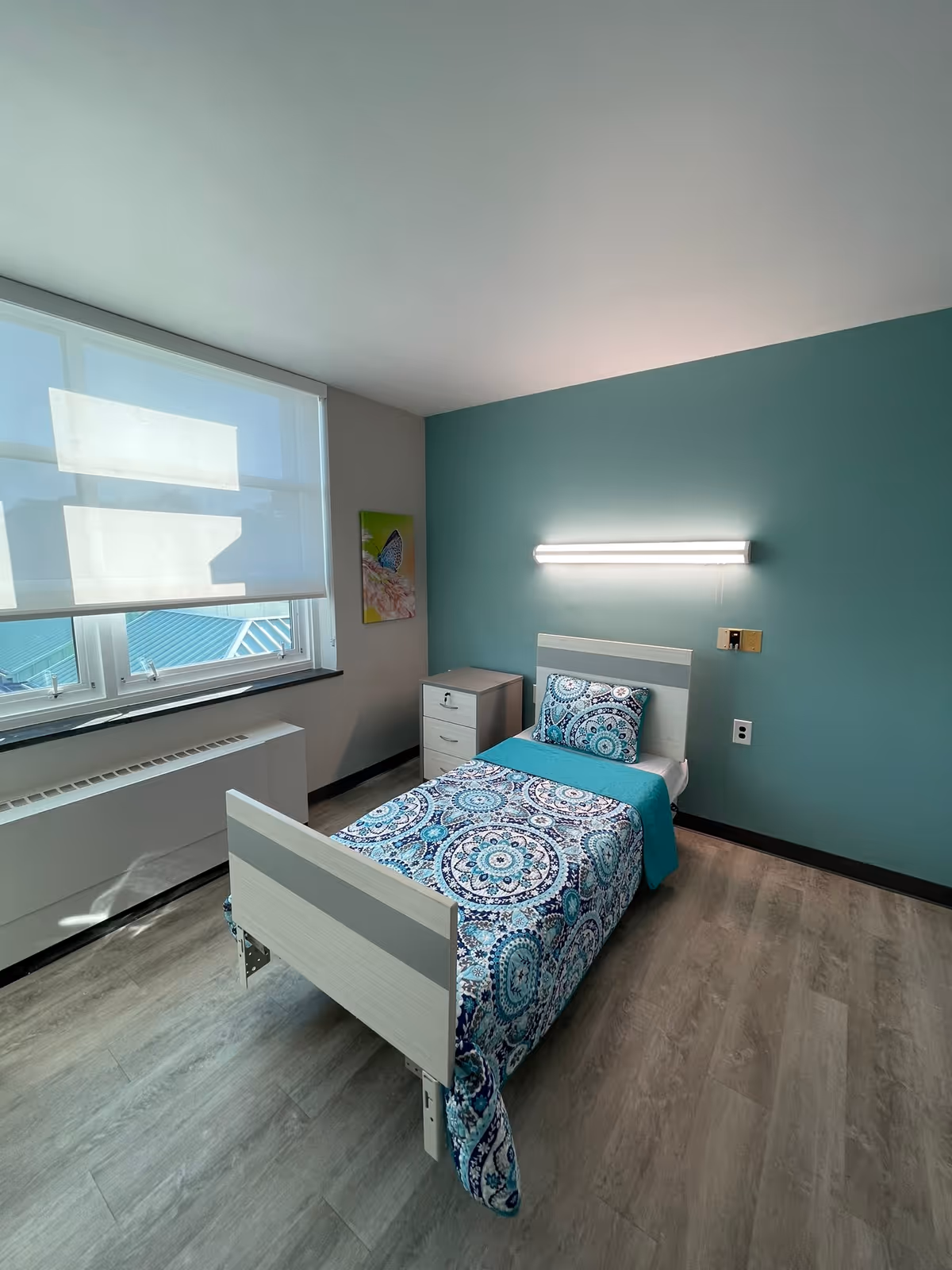 A single bed with a patterned blue and white bedspread and matching pillow in a room with teal and light gray walls. There is a small white nightstand next to the bed, a large window with white blinds, and a fluorescent light fixture above the bed. The floor is light wood laminate.