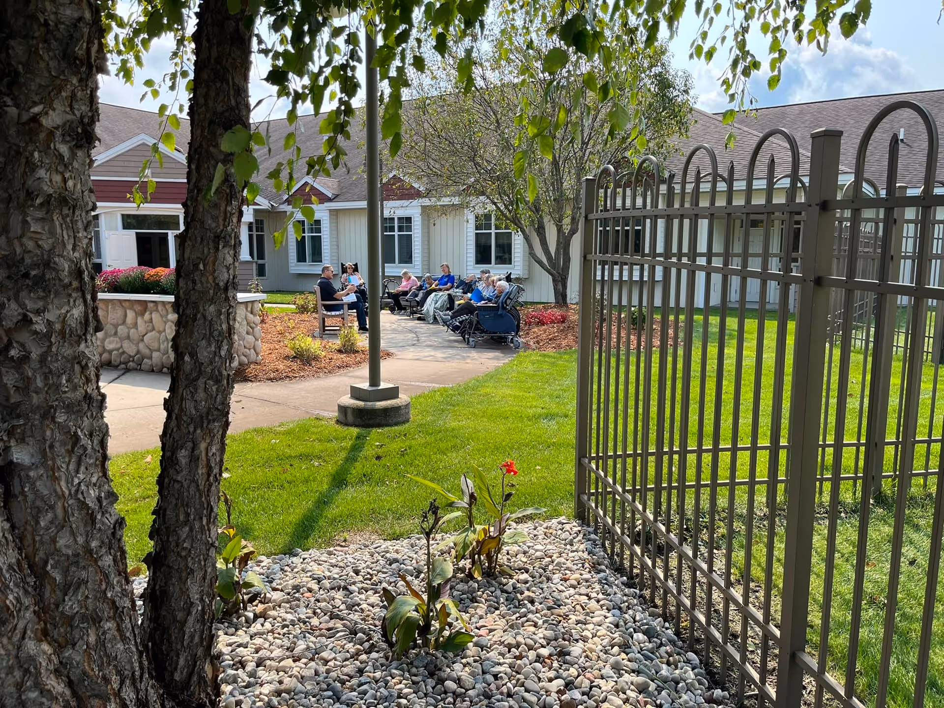 Outdoor courtyard area at Byron Manor with a group of elderly people sitting on chairs and wheelchairs along a paved walkway near a building. The scene includes green grass, a tree with hanging leaves, a stone planter with flowers, and a metal fence in the foreground.