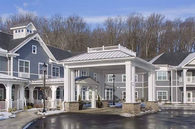 Exterior view of a senior living facility with gray siding and white trim, featuring a covered entrance with white columns and stone bases. The building is surrounded by a paved driveway and leafless trees in the background under a partly cloudy sky.