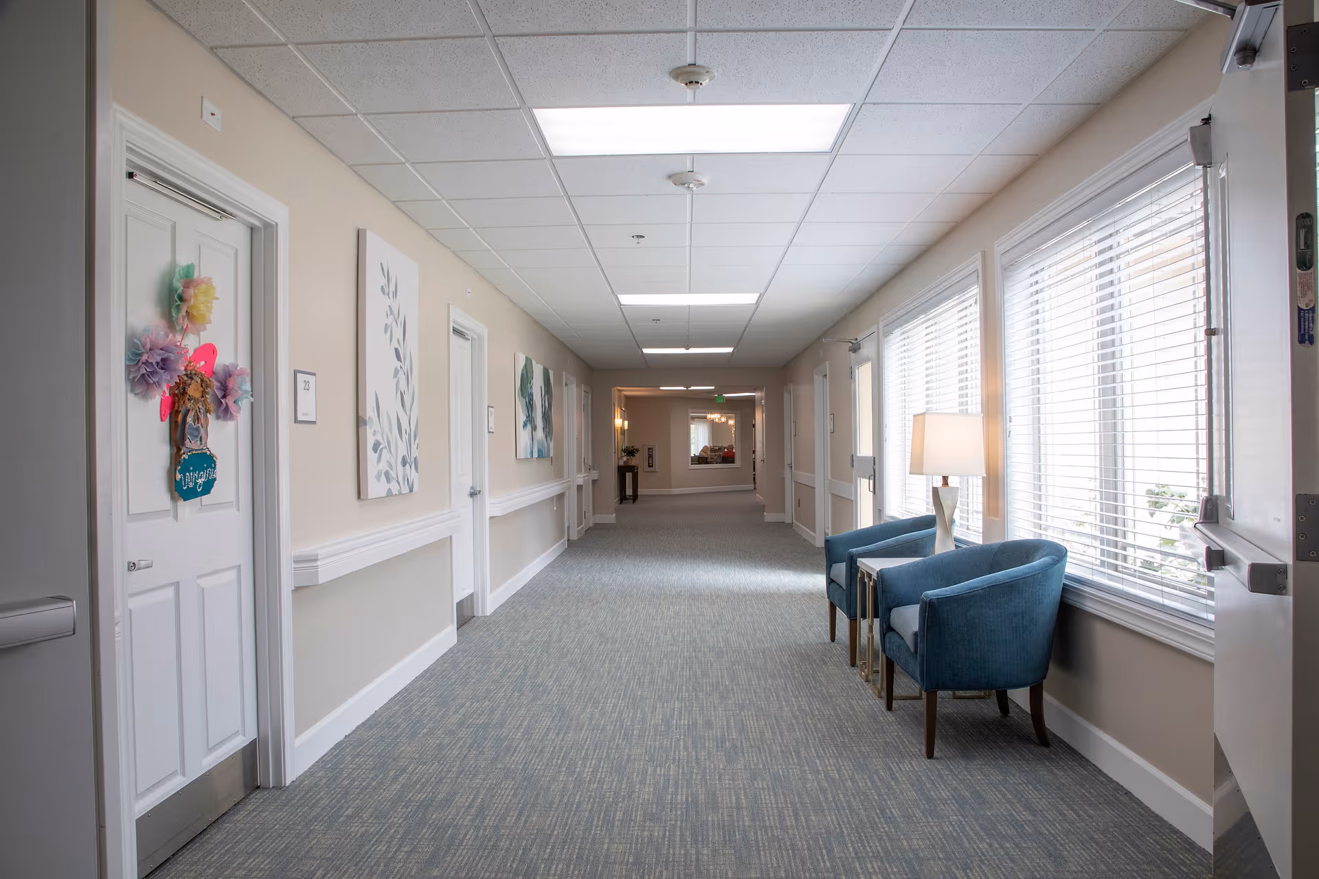 A well-lit hallway in a senior living facility with beige walls, carpeted floor, and white doors. One door is decorated with colorful paper flowers and a sign. The hallway has framed artwork on the walls and two blue armchairs with a small table and lamp near large windows with blinds on the right side.