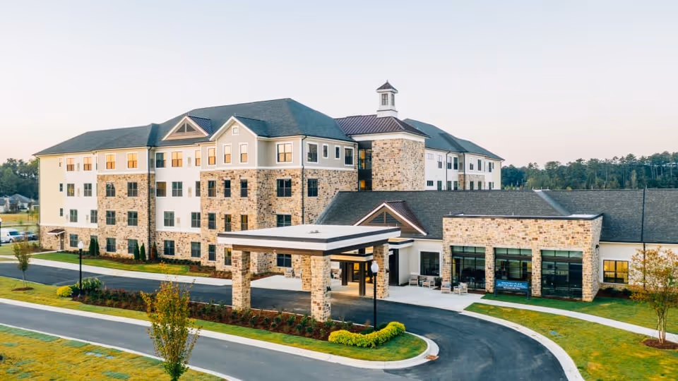 Exterior view of a large, multi-story senior living facility building with stone and beige siding, a covered entrance, and surrounding landscaped grounds with trees and a driveway.