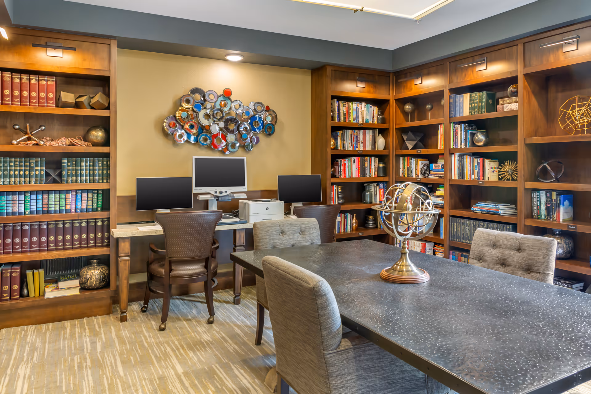 A well-lit room with wooden bookshelves filled with books and decorative items, a table with four upholstered chairs, and a desk with three computer monitors and a printer. A metallic globe sculpture is placed on the table, and a colorful abstract wall art hangs above the desk.